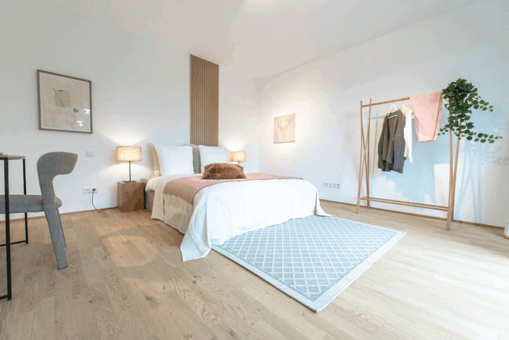 Hell eingerichtetes Schlafzimmer mit Doppelbett, zwei Nachttischlampen, Teppich und offener Kleiderstange mit Kleidung und Pflanze.