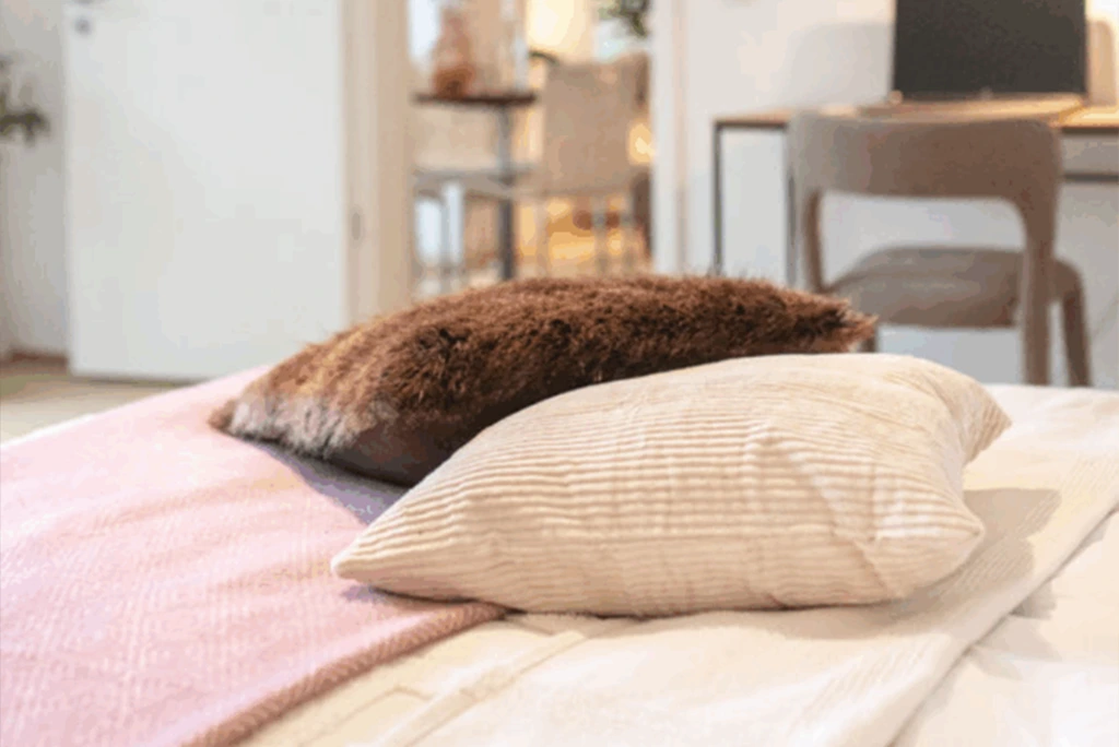 Zwei Kissen liegen auf einem Bett, eines braun und flauschig, das andere hell mit feinen Streifen.