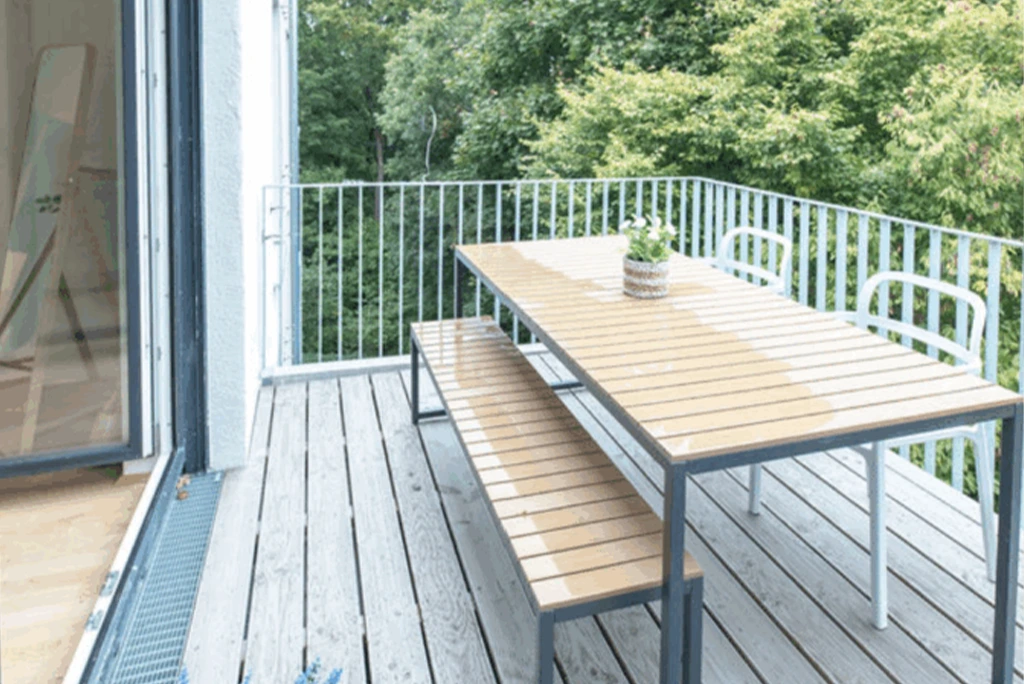Balkon mit grauem Holzboden, einem Tisch und einer Bank mit hellbraunen Holzplatten und grauem Metallgestell, zwei weißen Stühlen und einem kleinen Blumentopf auf dem Tisch.