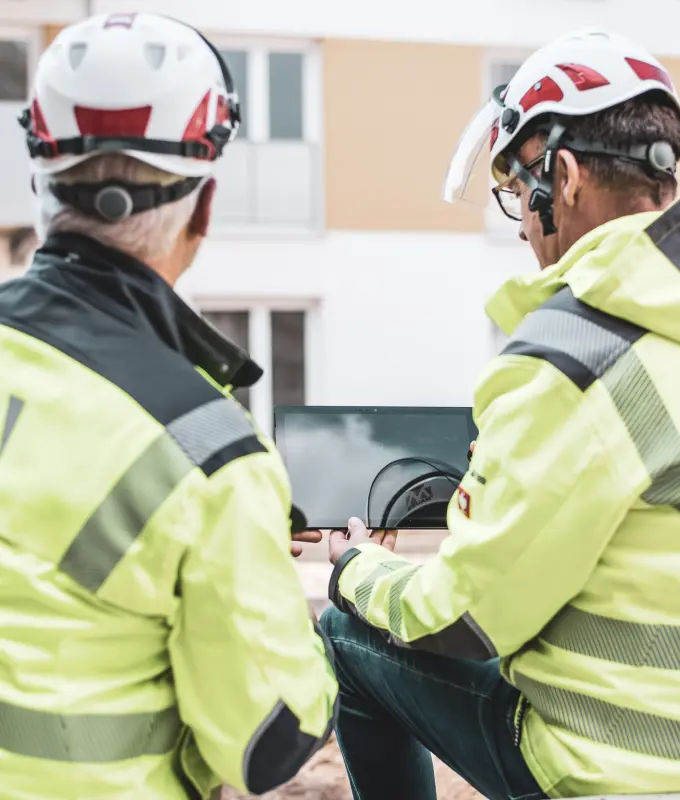 zwei Bauleiter blicken auf ein Tablet, im Hintergrund ein Gebäude
