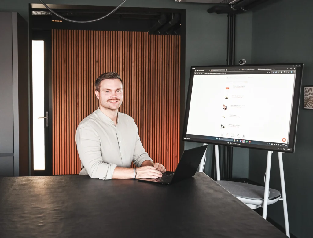 Person arbeitet mit Laptop am Tisch, digitales Whiteboard im Hintergrund