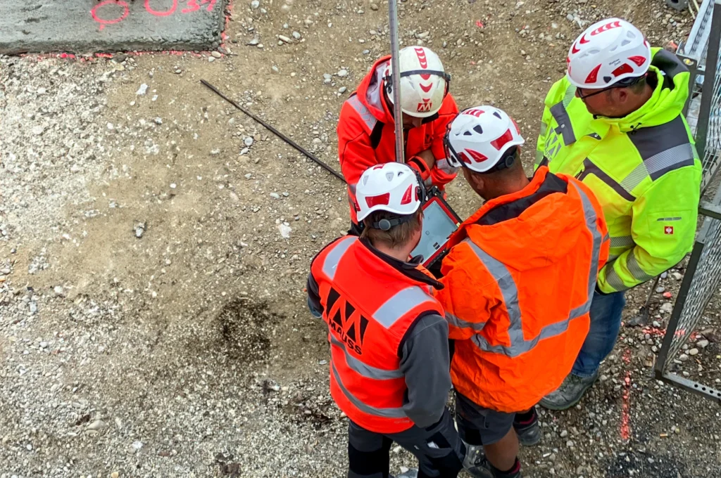 Gruppe von vier Mitarbeitern in Arbeitskleidung auf einer Baustelle, aus der Vogelperspektive fotografiert, beugen sich über ein Absteckgerät