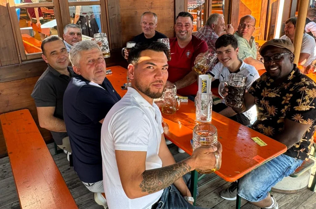 Gruppenfoto Kolleg*innen aus Ingolstadt, blicken lachend in die Kamera, sitzend an einer Biertischgarnitur, prosten mit Maßkrügen der Kamera zu, Volksfestatmosphäre