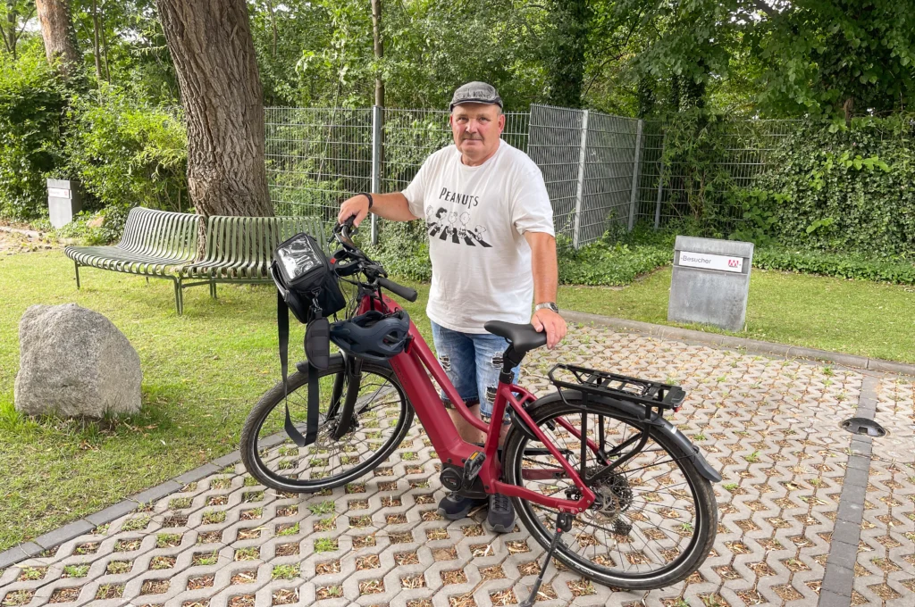 Mann mit Mütze steht neben einem dunkelroten E-Bike, im Hintergrund ist ein Parkplatz und eine grüne Parkanlage zu sehen