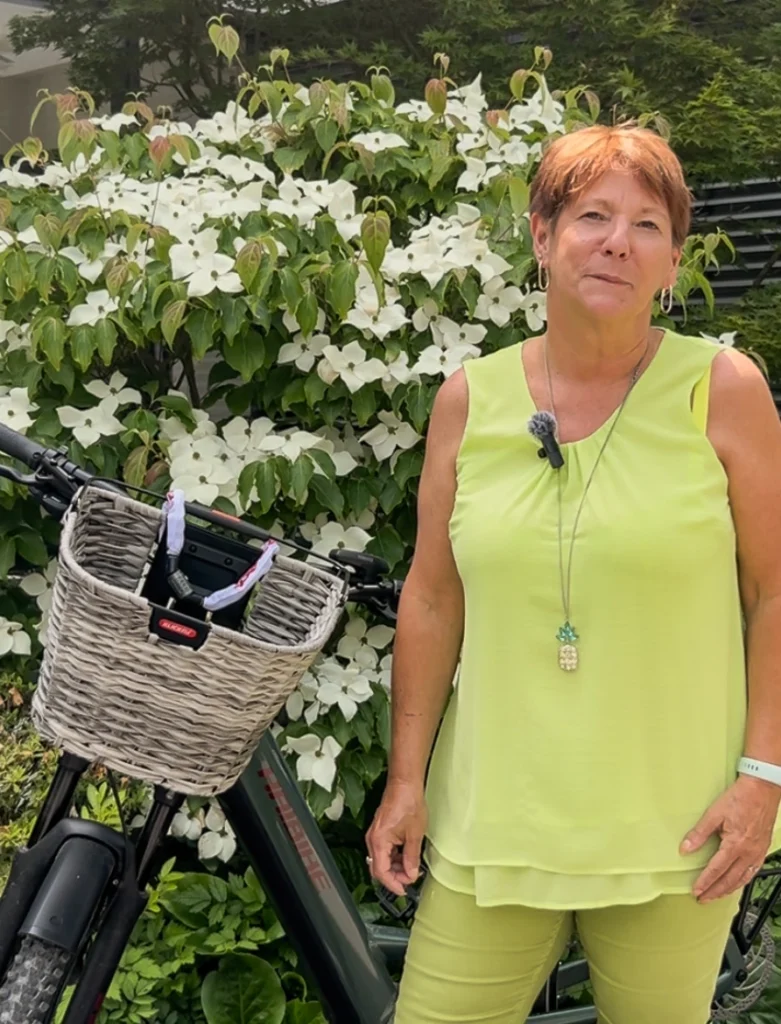 Frau in hellgrünem Outfit steht vor einem weiß blühenden Buch, ein Fahrrad mit Fahrradkorb am Lenker ist halb im Hintergrund zu sehen
