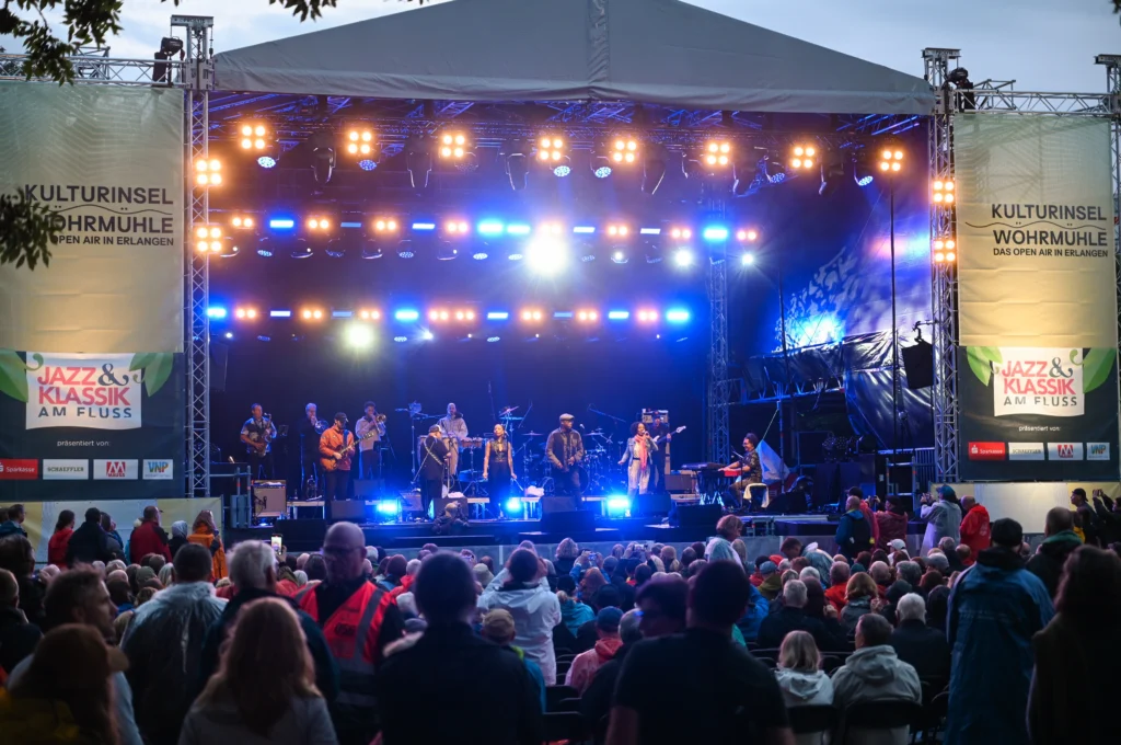 Frontalaufnahme Bühne Wöhrmühle 2025, Band auf der Bühne, blaue Beleuchtung, Abendstimmung, Besucher*innen sind von hinten zu sehen