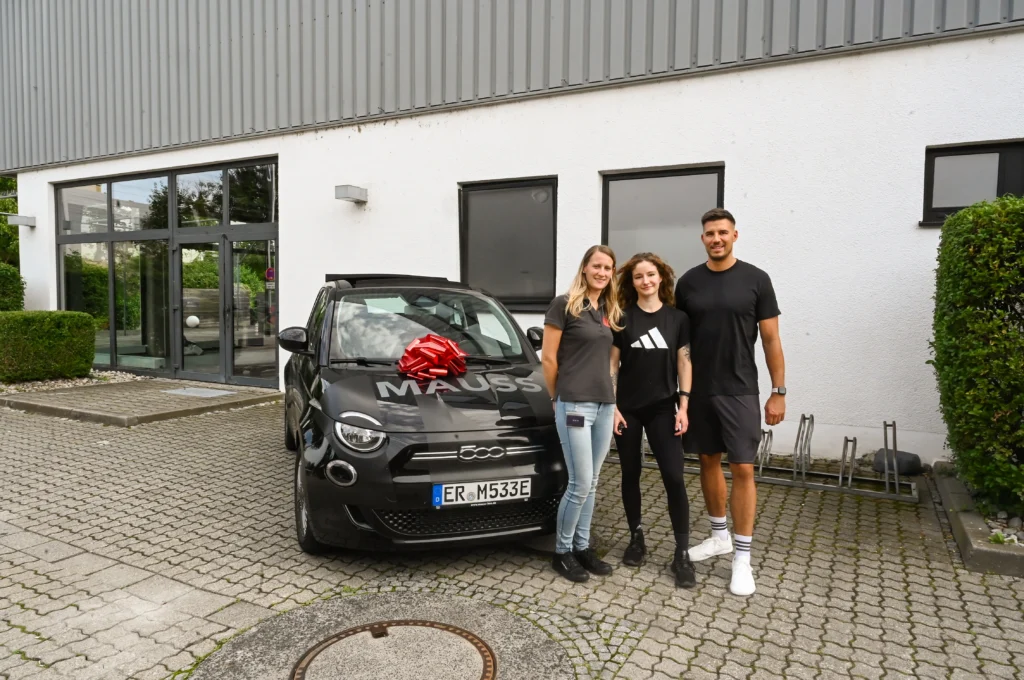 Drei lachende Personen stehen vor einem schwarzen Kleinwagen mit roter Schleife auf der Motohaube