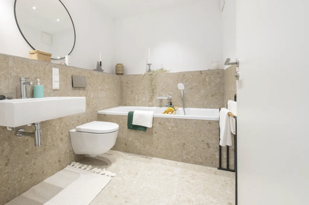 Helles Badezimmer mit wandhängendem WC, Badewanne mit Handbrause, rundem Spiegel und weißen Handtüchern.