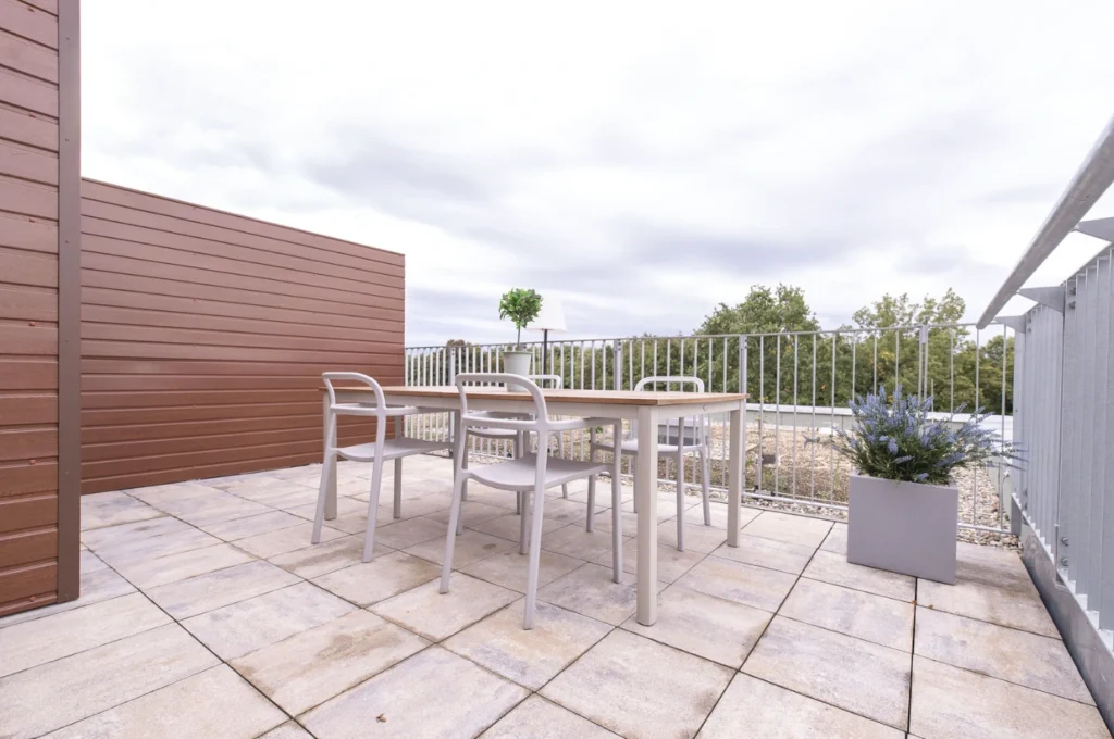 Terrasse mit quadratischem Tisch und vier weißen Stühlen, rechteckiger Blumentopf mit Lavendel, Metallgeländer und Holzwand.
