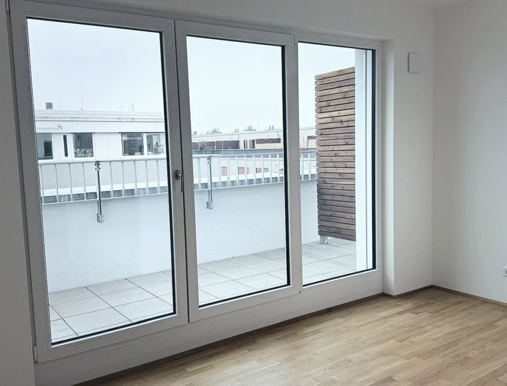 Helles, leeres Zimmer mit großen bodentiefen Fenstern, die auf einen Balkon mit weißer Brüstung und Holzverkleidung führen. Der Raum hat einen Holzfußboden.