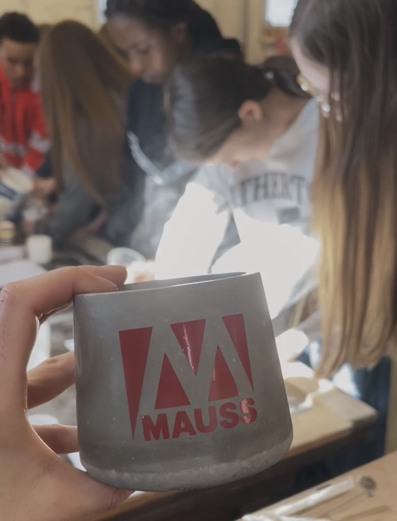 Das Bild zeigt im Vordergrund einem grauen Blumentopf mit unserem roten "MAUSS"-Logo, die von einer Hand gehalten wird. Im Hintergrund stehen mehrere Personen an einem Tisch und gießen Blumentöpfe aus Beton, wobei Formen und Materialien auf der Arbeitsfläche liegen.