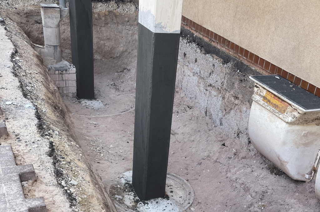 Das Bild zeigt eine schmale Baugrube entlang eines Gebäudes mit freigelegtem Erdreich und mehreren senkrechten Betonstützen auf runden Fundamenten. Die Betonstützen wurden im Rahmen einer Betoninstandsetzung saniert und sind schwarz versiegelt, um sie dauerhaft vor Feuchtigkeit und äußeren Einflüssen zu schützen.
