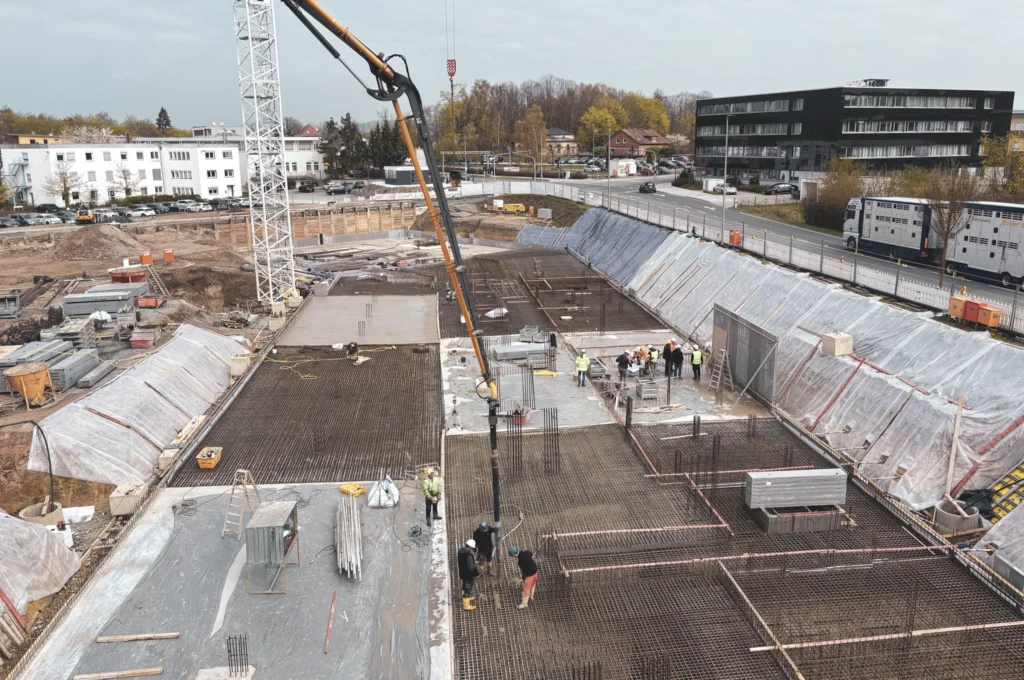 Das Bild zeigt eine große Baugrube, in der gerade die Bodenplatten aus Beton hergestellt werden. Mehrere Personen arbeiten auf dem vorbereiteten Untergrund, während ein langer Schlauch frischen Beton in die Baugrube fördert.