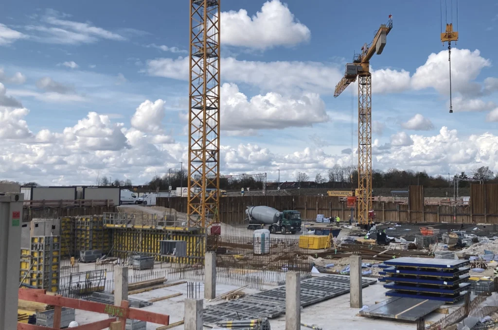 Das Bild zeigt eine aktive Baustelle mit zwei hohen Baukränen, Betonflächen, Schalungselementen und mehreren aus dem Boden ragenden Betonstützen. Im Hintergrund sind eine große Baugrube, Spundwände sowie Baustellenfahrzeuge und gelagertes Baumaterial zu sehen.