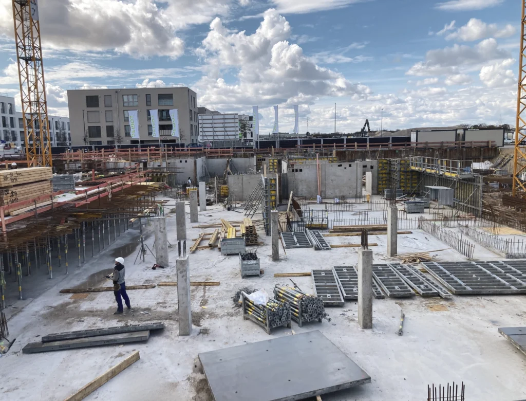 Das Bild zeigt eine weitläufige Baustelle mit zwei hohen Baukränen, Betonflächen, Schalungselementen und mehreren freistehenden Betonstützen. Zudem sind eine tiefe Baugrube, Baustellenfahrzeuge sowie gelagertes Baumaterial auf dem Areal zu erkennen.