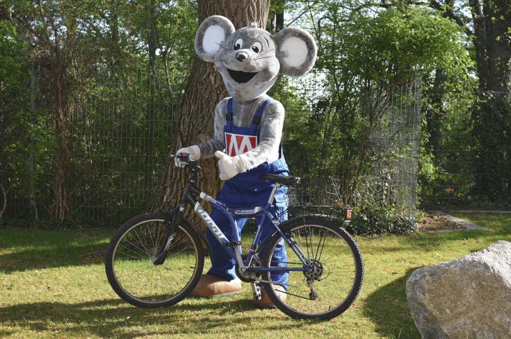 Das Bild zeigt das MAUS‑Maskottchen des Unternehmens, das auf einer Grünfläche mit einem Fahrrad vor einem Baum steht.