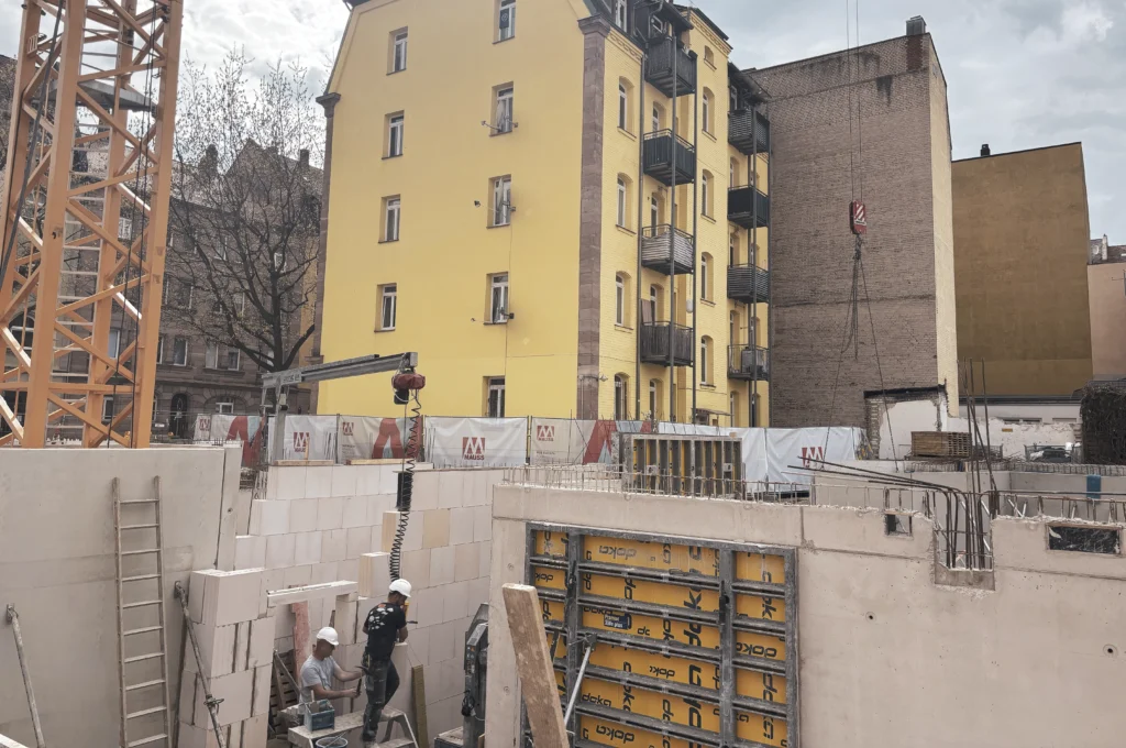 Das Bild zeigt den Rohbau mit gemauerten Wänden, einer großflächigen Schalung für eine Betonwand und mehreren arbeitenden Personen im Untergeschoss.