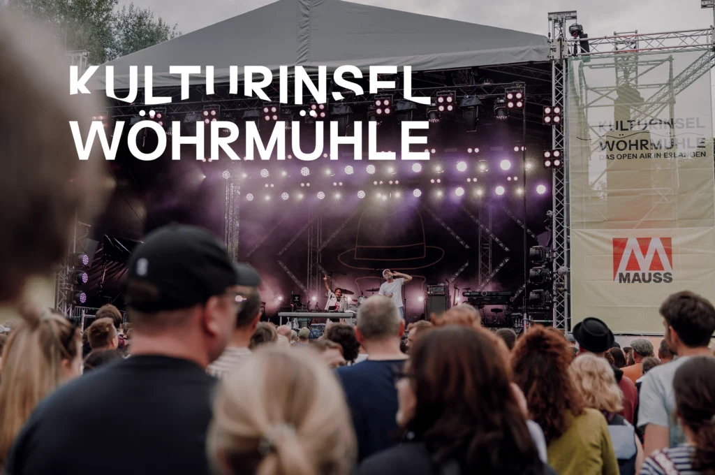 Das Bild zeigt eine große Open‑Air‑Bühne der Kulturinsel Wöhrmühle mit Beleuchtung und Live‑Band, davor steht ein dichtes Publikum. Seitlich ist ein Banner mit dem Schriftzug „Kulturinsel Wöhrmühle – Das Open Air in Erlangen“ sowie dem MAUSS‑Logo zu sehen.