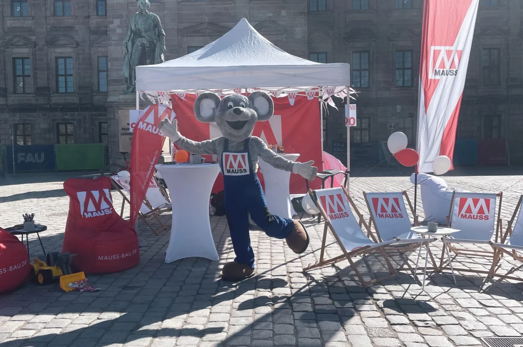 Das Bild zeigt das MAUSSkottchen vor dem MAUSS Stand bei der Rädli. Das Mausskottchen steht mit ausgebreiteten Armen auf einem gepflasterten Platz, umgeben von Liegestühlen, Fahnen und Sitzmöbeln mit MAUSS‑Logo sowie einem weißen Pavillon im Hintergrund.
