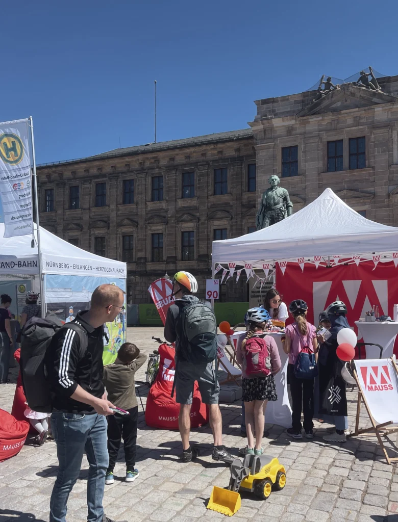 Das Bild zeigt unseren Stand bei der Rädli im Außenbereich mit einem weißen Pavillon und roten MAUSS-Logo‑Elementen wie Fahnen, Liegestühlen und Sitzsäcken. Mehrere Personen stehen davor, darunter auch Kinder mit Fahrradhelmen, während Spielmaterial und ein kleines Baustellenfahrzeug auf dem gepflasterten Boden liegen; im Hintergrund ist ein historisches Gebäude zu sehen.
