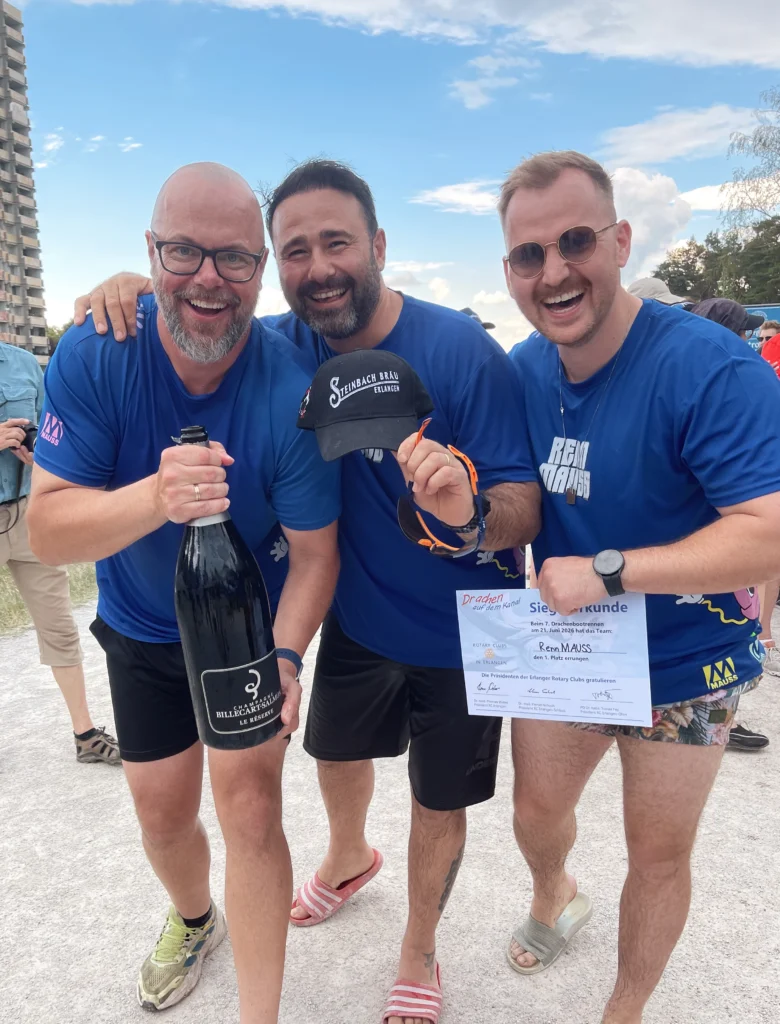 Drei Männer in blauen T-Shirts posieren im Freien, einer hält eine große Flasche, ein anderer eine Urkunde und eine schwarze Kappe.
