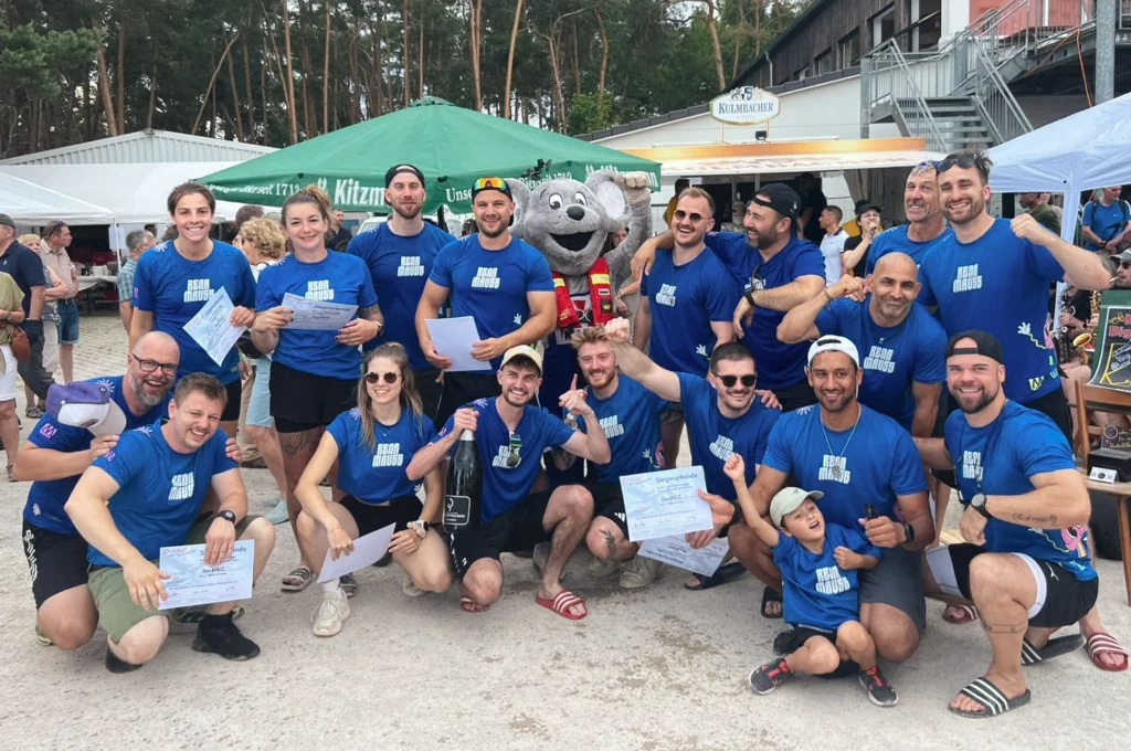 Das Bild zeigt eine große Gruppe von Personen, die im Außenbereich vor einem Biergarten zusammenstehen und blaue T‑Shirts tragen. Mehrere Personen halten Urkunden in den Händen, in der Mitte ist ein Maus‑Maskottchen mit erhobenen Armen zu sehen. Im Hintergrund stehen grüne Sonnenschirme, Verkaufsstände und Bäume, was auf eine Veranstaltung im Freien hinweist.