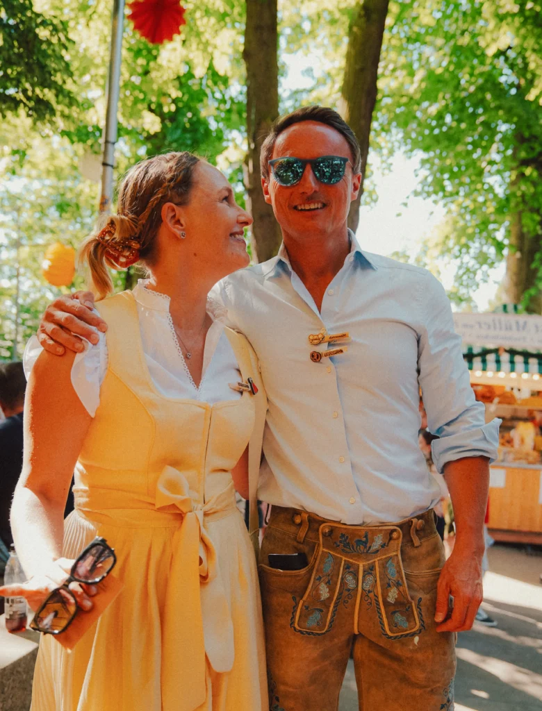Das Bild zeigt zwei Personen in traditioneller Tracht, die nebeneinander stehen und sich gegenseitig um die Schultern gelegt haben. Eine Person trägt ein gelbes Dirndl, die andere eine Lederhose mit hellem Hemd. Die Szene spielt im Außenbereich unter Bäumen, im Hintergrund sind Stände und Dekorationen eines Festes zu erkennen.