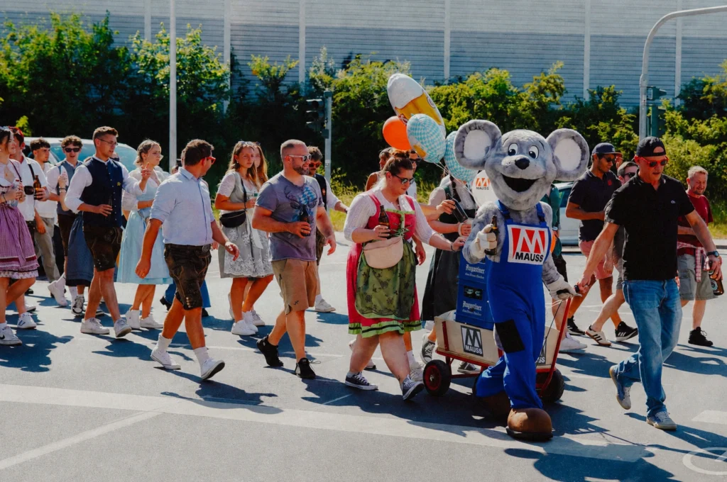 Das Bild zeigt Teilnehmende beim Kastenlauf zur Bergkirchweih, von denen viele traditionelle Tracht tragen. Begleitet werden sie vom MAUSS‑Maskottchen in einem Maus‑Kostüm mit blauem Overall, das einen Handwagen zieht.