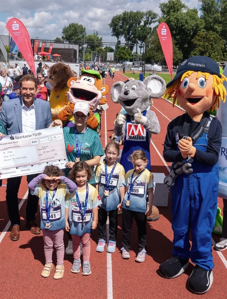 Das Bild zeigt eine Gruppe von Kindern und Erwachsenen auf einer Laufbahn bei einer Sportveranstaltung. Im Vordergrund stehen mehrere kostümierte Figuren, darunter ein Maus‑Maskottchen sowie weitere Tier‑ und Figurenkostüme, neben Kindern mit Startnummern und Medaillen. Im Hintergrund sind Zuschauende, rote Fahnen mit Logos und die Sportanlage zu sehen.
