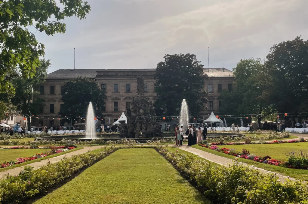 Das Bild zeigt eine symmetrisch angelegte Gartenanlage mit Rasenflächen, Blumenbeeten und geraden Wegen. In der Mitte befindet sich ein Brunnen mit Wasserspielen vor einem historischen Gebäude, davor sind einzelne Personen und Pavillons zu sehen.