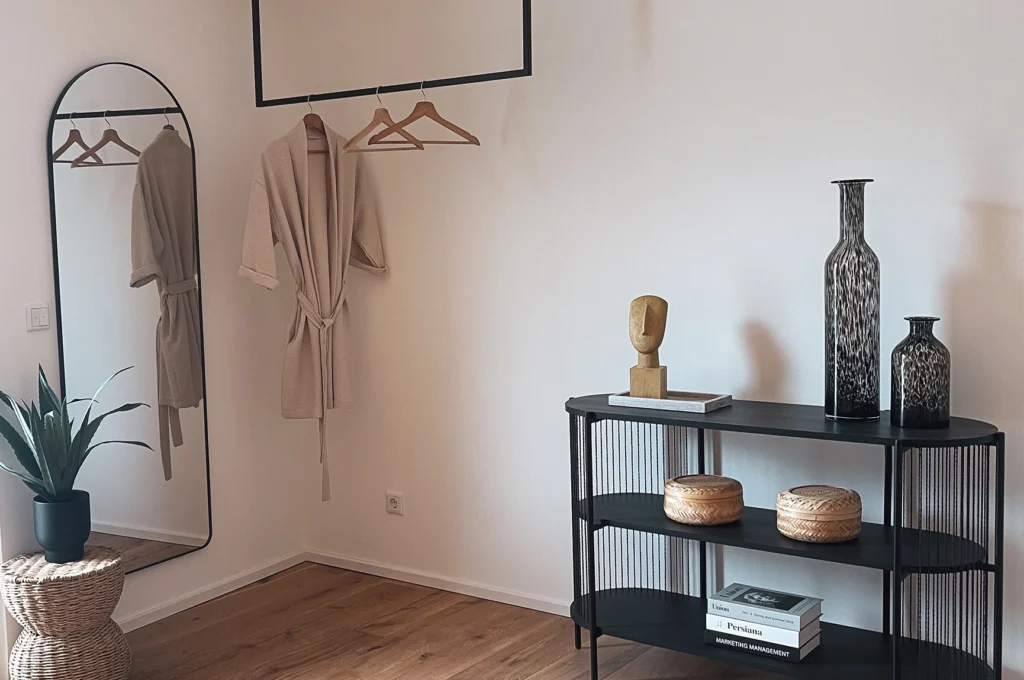 Das Bild zeigt einen hellen Innenraum mit Holzboden, einer offenen Kleiderstange mit Bademantel und einem großen Standspiegel. Daneben steht ein schmales Regal mit Vasen, Körben und Büchern sowie eine Topfpflanze auf einem gewebten Beistelltisch.