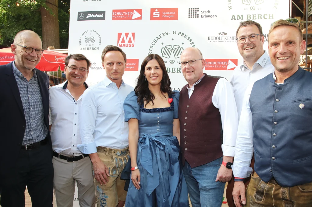 Das Bild zeigt eine Gruppe von Personen, die dicht nebeneinander vor einer Sponsoren‑ und Logo‑Wand stehen. Viele tragen traditionelle Tracht, darunter Lederhosen, Westen und ein Dirndl. Im Hintergrund sind verschiedene Logos und Schriftzüge auf einem großen Banner zu sehen.