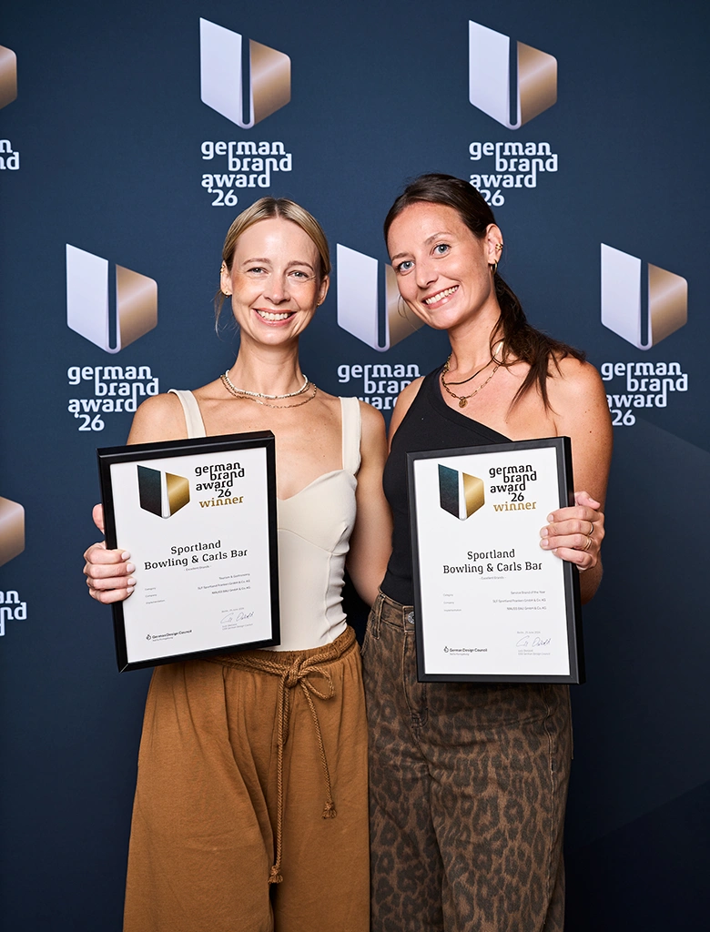 Franziska Groß und Kristina Schlerf stehen vor einer Wand mit dem Schriftzug 'german brand award 26' und halten jeweils eine Urkunde mit dem Titel 'german brand award winner' für 'Sportland Bowling & Cats Bar'