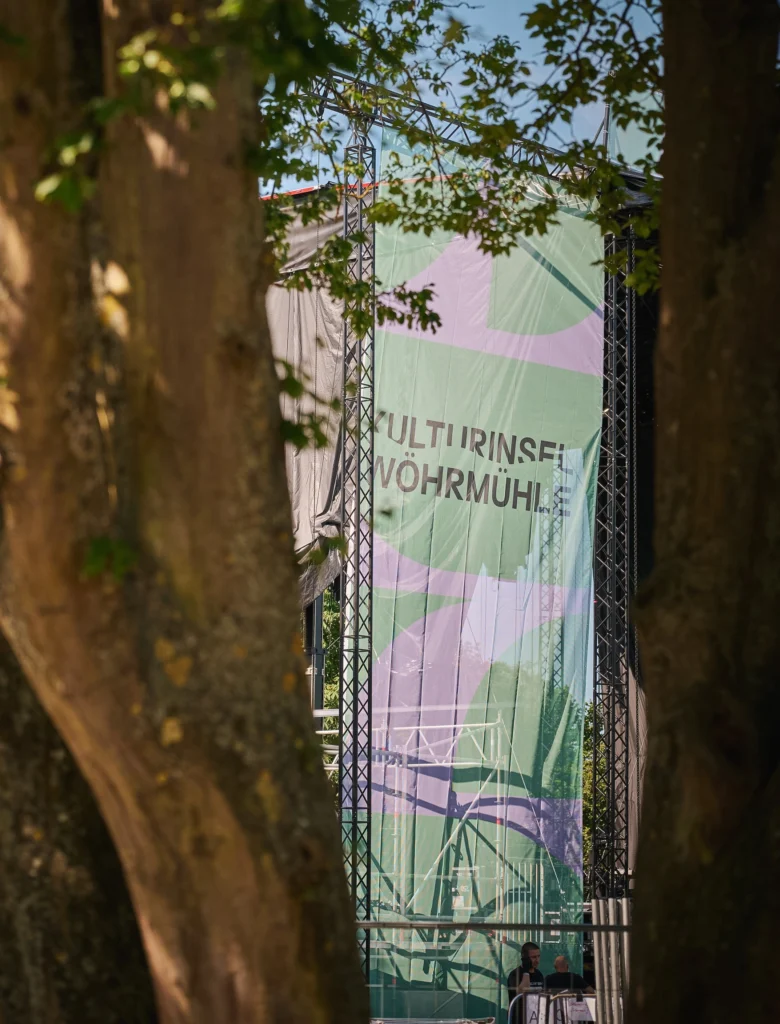Blick durch Baumstämme auf ein großes Banner mit der Aufschrift 'KULTURINSEL WÖHRMÜHLE'