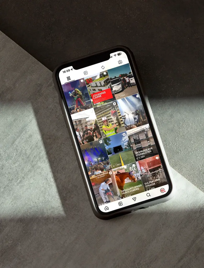 Ein Smartphone mit abgerundeten Ecken liegt auf einer Oberfläche, die aus zwei unterschiedlich strukturierten Materialien besteht. Das Display zeigt eine Social-Media-App mit einer Rasteransicht aus mehreren quadratischen Vorschaubildern, die verschiedene Motive und Farben zeigen. In der oberen Leiste sind Symbole für Navigation und Benachrichtigungen zu sehen. Das Gerät wirft einen Schatten auf die Oberfläche.