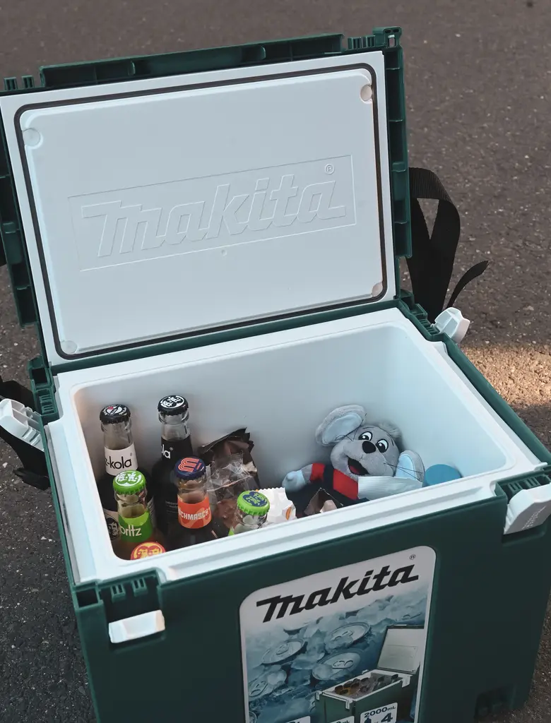 Eine grüne Kühlbox mit weißem Deckel steht geöffnet auf Asphalt. Im Inneren liegen mehrere Glasflaschen mit unterschiedlichen Etiketten, eine kleine Tüte mit Snacks und ein graues Stofftier in Elefantenform. Auf dem Deckel und an der Vorderseite ist jeweils ein Makita-Logo zu sehen.