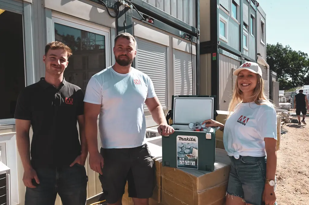Drei Personen stehen nebeneinander vor einem Baucontainer. Alle tragen helle Oberteile mit einem Firmenlogo auf der linken Brust. In der Mitte steht eine grüne Kühlbox mit geöffnetem Deckel, darin sind mehrere Getränkedosen sichtbar. Die Personen lächeln und blicken in Richtung Kamera. Im Hintergrund sind weitere Container und Baumaterialien zu erkennen.