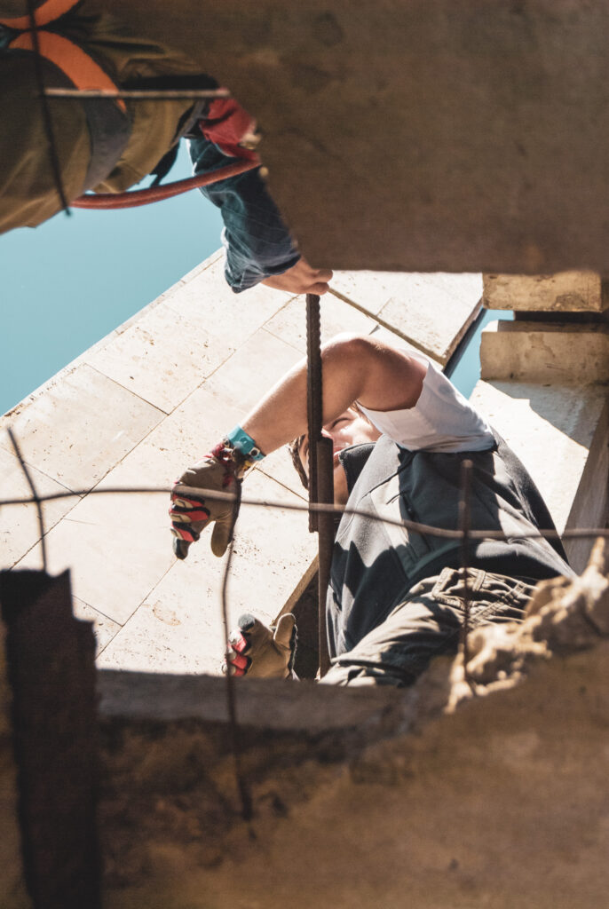 Das Bild zeigt eine Person auf einer Baustelle, die einen Stahlstab hält. Die Aufnahme erfolgt durch eine Öffnung in einer Betonwand, umgeben von Bewehrungsstäben und Betonkanten.