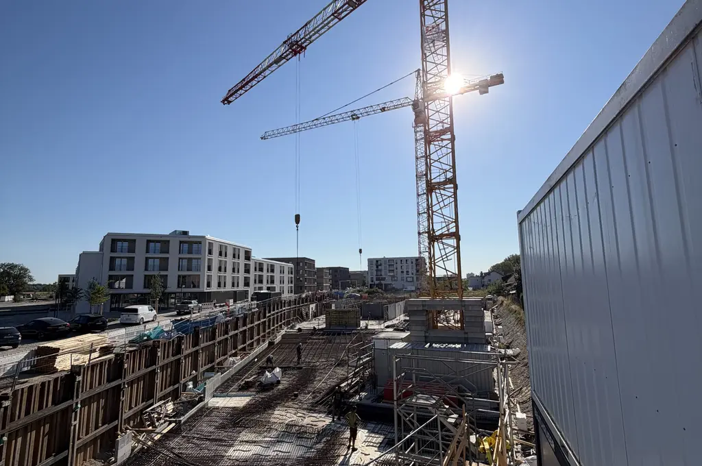 Großbaustelle bei Tageslicht mit zwei hohen Baukränen im Vordergrund. Links stehen moderne, mehrstöckige Wohngebäude, rechts ein weißer Container. Im Baustellenbereich sind zahlreiche Baumaterialien, Gerüste und Absperrungen zu sehen. Der Himmel ist wolkenlos und die Sonne scheint von rechts oben ins Bild.