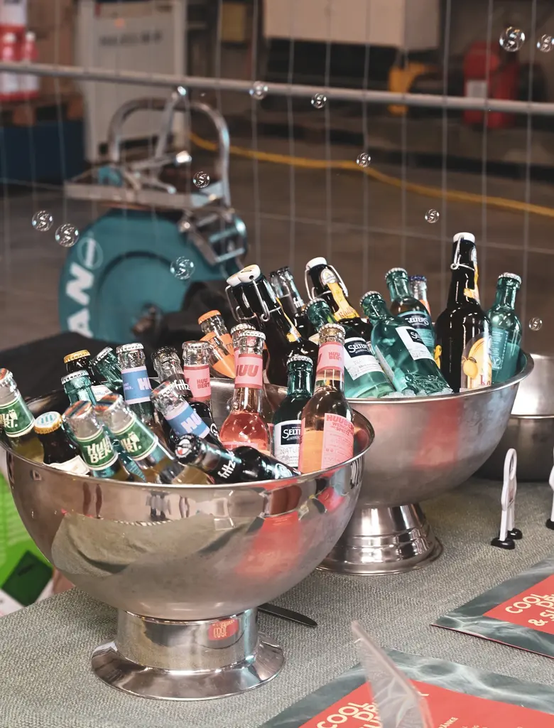 Zwei große, silberne Schalen stehen nebeneinander auf einem Tisch und sind mit verschiedenen Flaschen gefüllt. Die Flaschen enthalten unterschiedliche Getränke, darunter Wasser und Limonade, und sind in Eis gekühlt. Im Vordergrund liegen mehrere Broschüren oder Flyer auf dem Tisch. Im Hintergrund sind industrielle Geräte und eine Absperrung zu sehen. Einige Seifenblasen schweben durch das Bild.