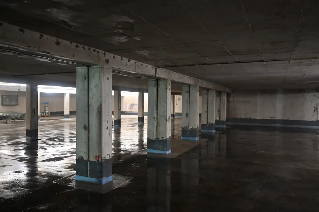Mehrere Betonpfeiler stehen in einem weitläufigen, leeren Raum mit niedriger Decke. Der Boden ist dunkel und glänzend, als wäre er feucht. Die Wände und Pfeiler zeigen Abnutzungsspuren und Verfärbungen. Das Licht im Raum ist diffus und stammt von Leuchtstoffröhren entlang der Decke.