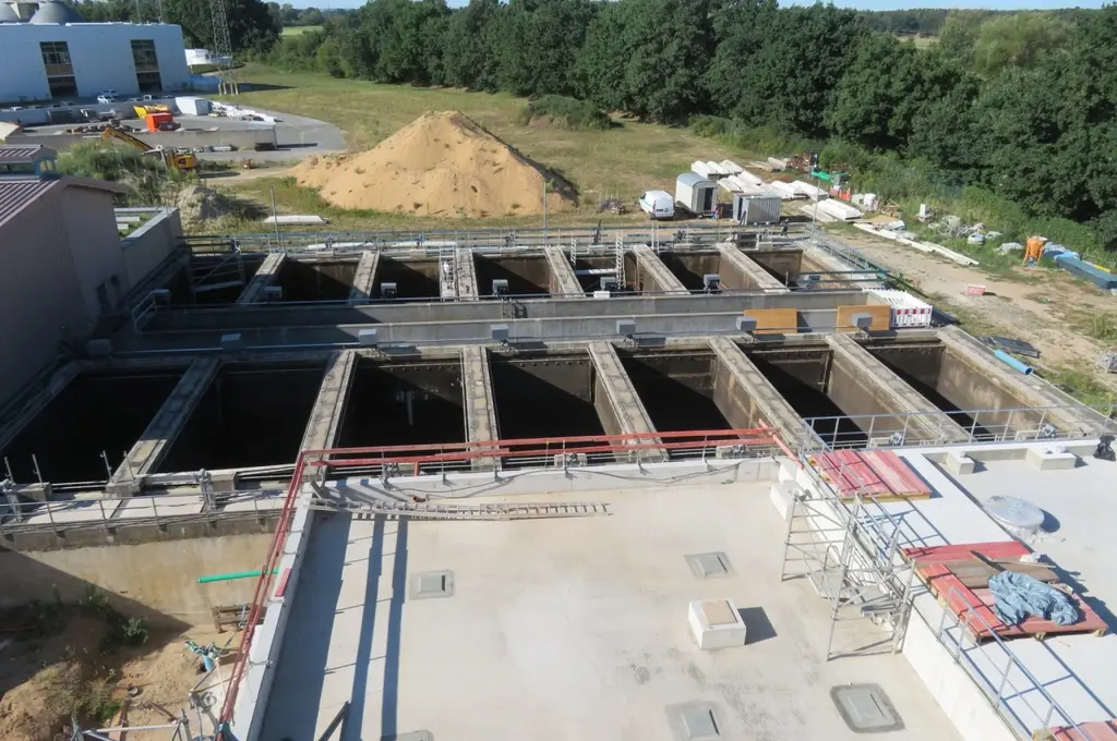 Im Vordergrund befindet sich eine große, rechteckige Betonfläche mit mehreren quadratischen Abdeckungen und Gerüsten. Dahinter sind parallele, offene Becken mit dunklem Wasser angeordnet, die durch Betonwände getrennt sind. Im Hintergrund stehen Bäume, eine Sandaufschüttung und verschiedene Baucontainer sowie Fahrzeuge auf einer Wiese. Die Szene zeigt eine industrielle Anlage im Freien, vermutlich eine Kläranlage.