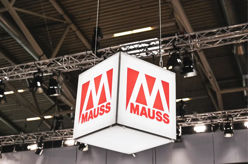 Im Mittelpunkt hängt ein würfelförmiges Leuchtelement von der Decke, dessen Seitenflächen jeweils das rote Logo mit dem Schriftzug 'MAUSS' zeigen. Die Konstruktion ist von Metallträgern und Scheinwerfern umgeben, die Teil einer Messehalle sind. Im Hintergrund sind weitere Beleuchtungselemente und die Decke der Halle sichtbar.