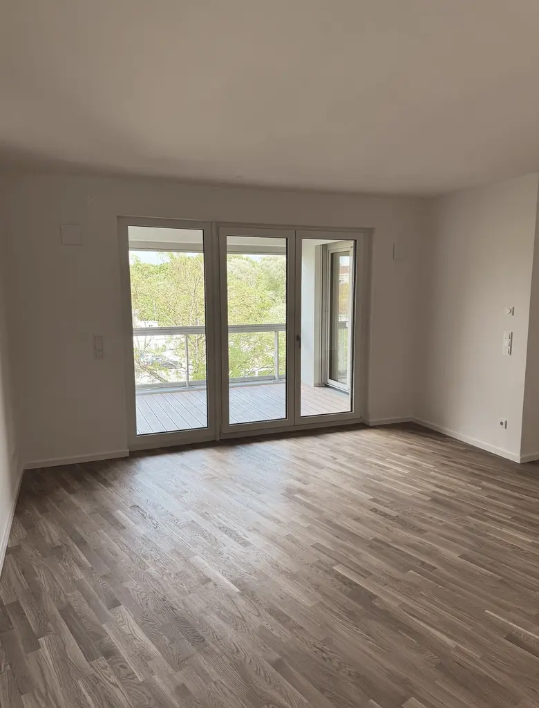 Heller Raum mit großem Fenster und Glastür, die auf einen Balkon führen. Der Boden besteht aus Holz mit deutlicher Maserung. Die Wände sind weiß gestrichen, der Raum ist leer und wirkt modern und gepflegt. Draußen sind grüne Bäume und ein Teil des Balkongeländers sichtbar.