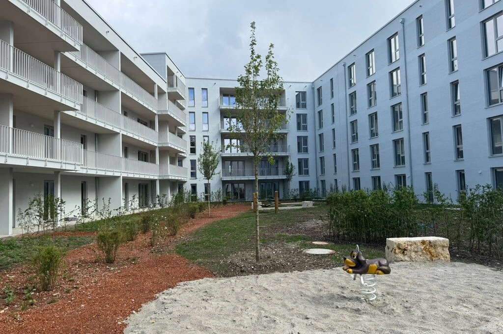 Innenhof einer modernen Wohnanlage mit viergeschossigen, hellen Gebäuden. Die Fassaden sind glatt und hellblau gestrichen, mit durchgehenden Balkonen und großen Fenstern. Im Zentrum des Hofs steht ein einzelnes junges Bäumchen, umgeben von niedrigen Sträuchern und Beeten mit rotem Mulch. Im Vordergrund befindet sich ein Sandspielbereich mit einer Wipptier-Federwippe in Hundeform und einem großen Naturstein. Der Himmel ist bewölkt.