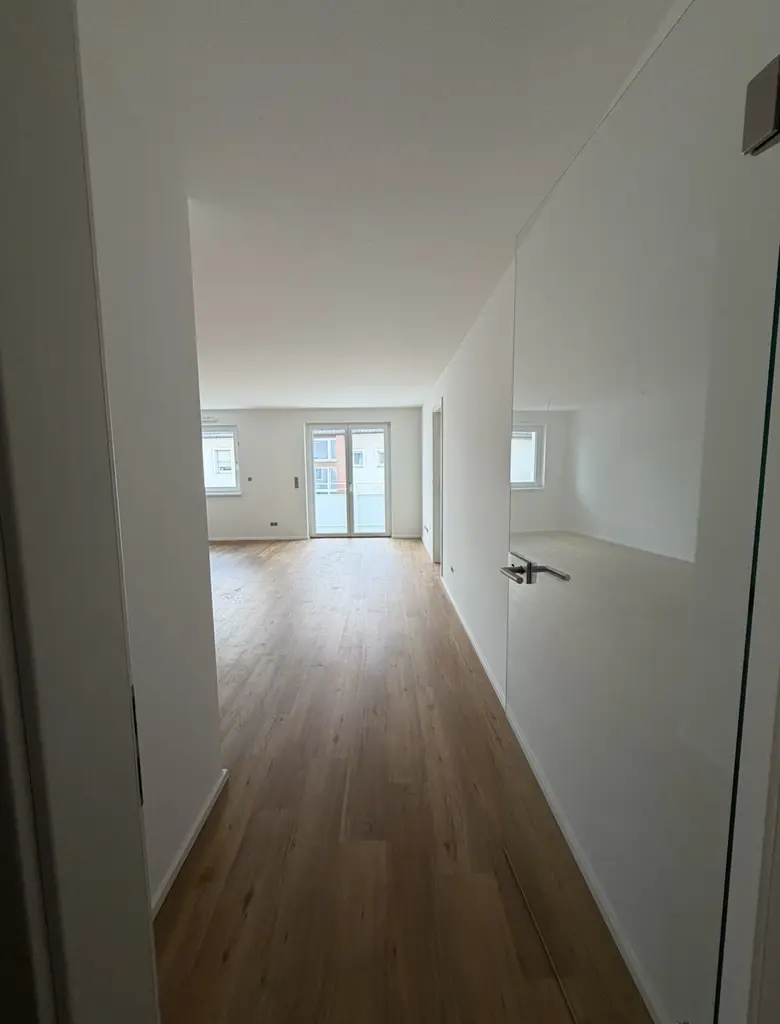 Heller Raum mit glattem Holzboden und weißen Wänden. Im Vordergrund eine geöffnete Tür mit silbernem Griff, dahinter ein großzügiger Wohnbereich mit mehreren Fenstern und einer Glas-Doppeltür, die auf einen Balkon oder eine Terrasse führt. Die Fenster lassen viel Tageslicht herein.
