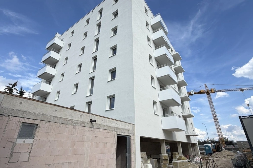 Ein mehrstöckiges Wohngebäude mit weißer Fassade steht im Vordergrund. Mehrere Balkone sind an der rechten Seite angeordnet. Im unteren Bereich des Gebäudes befinden sich offene Flächen und Baumaterialien. Links ist eine niedrige Mauer aus hellen Steinen zu sehen. Im Hintergrund ragt ein gelber Baukran in den Himmel, daneben sind weitere Baustellenmaterialien und ein Schild sichtbar. Der Himmel ist blau mit einigen weißen Wolken.