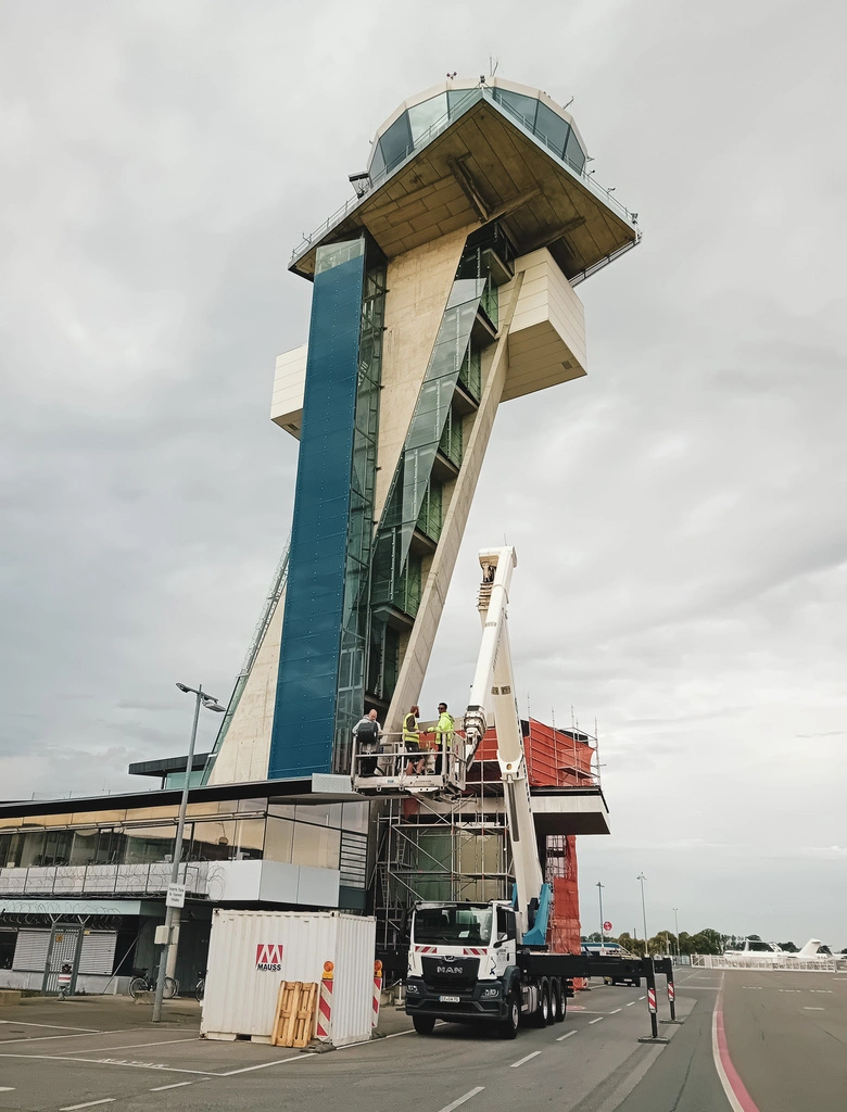 Ein markanter, schräg aufragender Tower mit verglaster Kanzel steht auf einem Flughafengelände. Vor dem Gebäude befindet sich ein Hubsteiger, auf dessen Plattform mehrere Personen in Warnwesten arbeiten. Das Wetter ist bewölkt, der Boden asphaltiert. Im Vordergrund sind Absperrungen und ein weißer Container zu sehen.