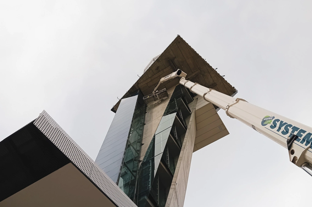 Ein Hubsteiger mit ausfahrbarem Arm reicht bis zu einer modernen Gebäudefassade aus Glas und Beton. Eine Person steht im Arbeitskorb und führt Arbeiten an der Unterseite eines weit auskragenden Flachdachs aus. Die Perspektive ist von unten nach oben gerichtet, der Himmel ist bewölkt.