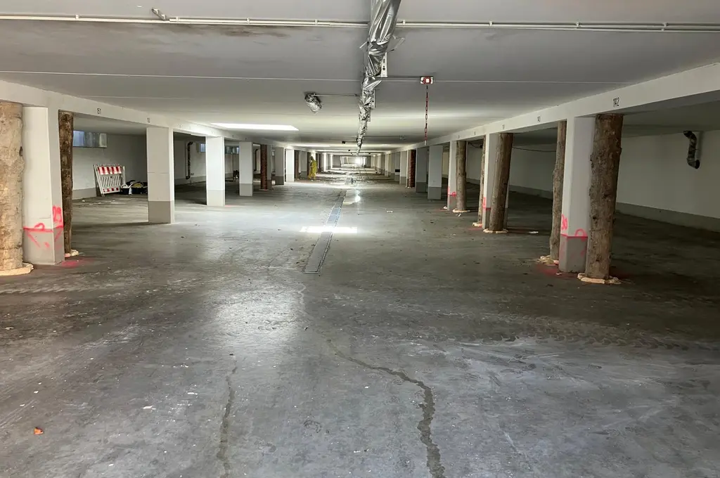 Betonpfeiler mit auffälligen rosa Markierungen stehen in regelmäßigen Abständen in einer langgestreckten Tiefgarage. Die Decke ist niedrig, das Licht gleichmäßig und die Fläche wirkt leer. Im Hintergrund sind Absperrgitter zu erkennen.